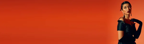 Charmante et gracieuse femme asiatique en tenue de printemps élégante regardant loin sur fond rouge, cheveux bruns, maquillage audacieux, gants longs, robe noire, concept de printemps à la mode, bannière — Photo de stock
