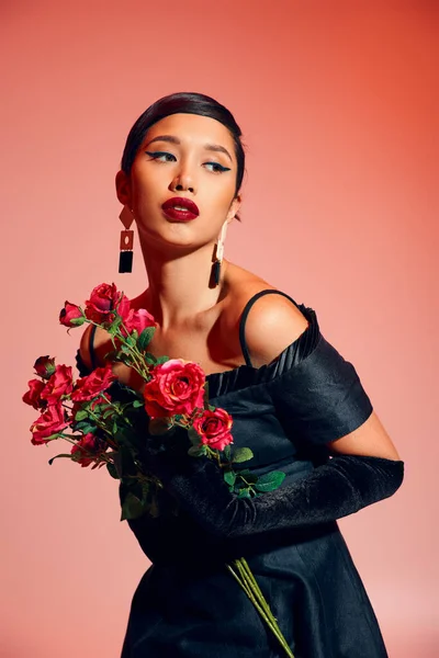 Young and expressive asian woman with brunette hair, bold makeup, in black cocktail dress, long gloves and trendy earrings holding roses and looking away on pink background, stylish spring concept — Stock Photo