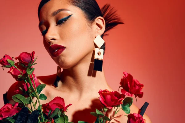 Portrait of attractive and fashionable asian woman with brunette hair, bold makeup, trendy hairstyle and earrings posing with red and fresh roses on pink and red background, spring fashion concept — Stock Photo