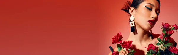 Retrato de mujer asiática bonita y sensual con cabello moreno, maquillaje audaz, pendientes de moda y peinado posando cerca de rosas frescas sobre fondo rosa y rojo, moda gen z, primavera con estilo, bandera - foto de stock