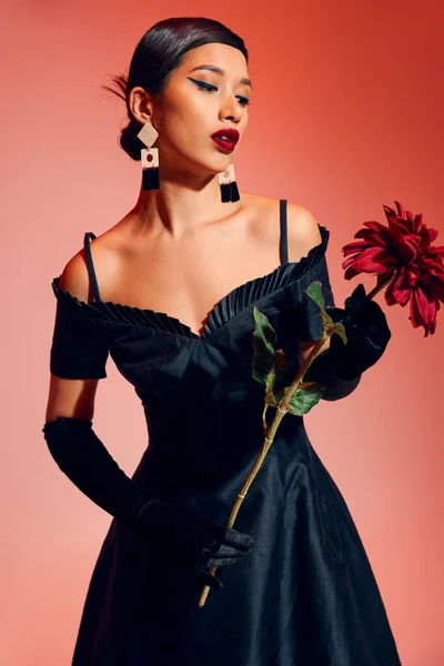 Mujer asiática joven y de moda con maquillaje audaz, en guantes largos negros y vestido de cóctel mirando peonía borgoña mientras está de pie sobre fondo rojo y rosa, fotografía de moda de primavera - foto de stock