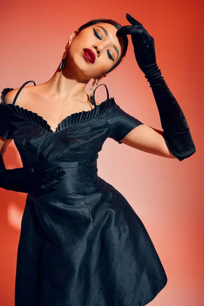 Mujer asiática joven y seductora con maquillaje audaz, cabello moreno, en guantes largos negros y vestido de cóctel posando con los ojos cerrados sobre fondo rojo y rosa, cultura juvenil, concepto de primavera de moda - foto de stock