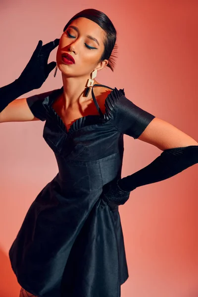 Femme asiatique hypnotisante avec un maquillage audacieux, cheveux bruns, en gants longs et robe de cocktail noire debout dans une pose élégante avec les yeux fermés sur fond rouge et rose, concept de mode de printemps — Photo de stock