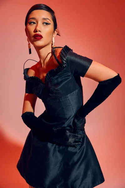 Expressive and fashionable asian woman in trendy earrings, long gloves and black cocktail dress standing with hands on hip and looking away on red and pink background, spring fashion, generation z — Stock Photo