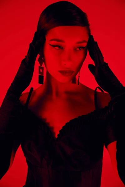 Retrato de encantadora mujer asiática con maquillaje audaz, en pendientes de moda, guantes largos y vestido de cóctel negro tocando la cabeza sobre un fondo vibrante con efecto de iluminación roja, elegante concepto de primavera - foto de stock