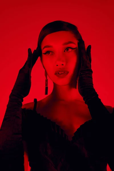Retrato de impresionante mujer asiática con cabello moreno y maquillaje audaz cogidas de la mano cerca de la cara y mirando hacia otro lado sobre un fondo vibrante con efecto de iluminación roja, vestido negro, guantes largos, primavera de moda - foto de stock