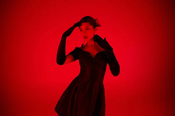 Молодая азиатка в черных длинных перчатках и элегантном коктейльном платье, стоящая в выразительной позе и глядя в сторону на ярком фоне с эффектом красного освещения, молодежной культуры, модной весны — стоковое фото