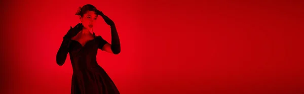 Young and fashionable asian woman in black cocktail dress and long gloves posing on vibrant background with red lighting effect and copy space, spring fashion photography, banner — Stock Photo
