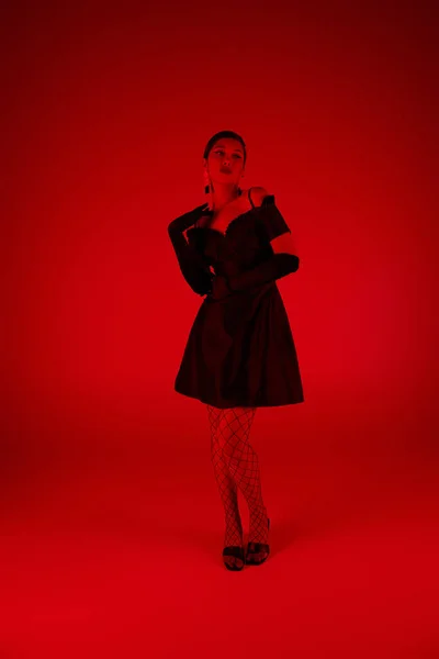 Весенняя модная фотография, полная длина стильной азиатской женщины в черном коктейльном платье, трико и длинные перчатки, стоящие на ярком фоне с эффектом красного освещения, поколение z — стоковое фото