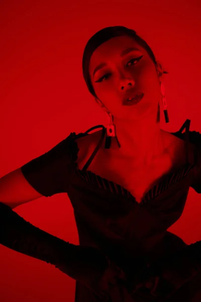 Ген z мода, молодая азиатская женщина в черном коктейльном платье, модные серьги и длинные перчатки, глядя на камеру с соблазнительным взглядом на ярком фоне с эффектом красного освещения, стильная весенняя концепция — стоковое фото