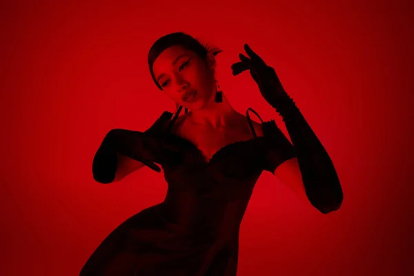Експресивна азіатська жінка з брюнеткою волосся, в чорних довгих рукавицях і стильна коктейльна сукня позує на яскравому фоні з ефектом червоного освітлення, модною весною, концепцією молодіжної культури — стокове фото
