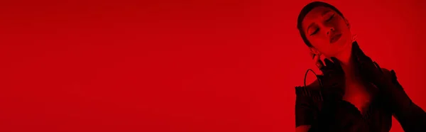 Seductora mujer asiática en vestido elegante negro y guantes largos tocando el cuello mientras posa con los ojos cerrados sobre un fondo vibrante con efecto de iluminación roja, cultura juvenil, elegante concepto de primavera, pancarta - foto de stock