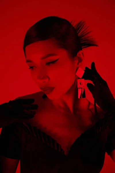 Портрет чарівної азіатки з брюнеткою волоссям і сміливим макіяжем, в чорній сукні і довгих рукавичках, що торкаються модного сережки на яскравому фоні з ефектом червоного освітлення, концепція весняної моди — стокове фото