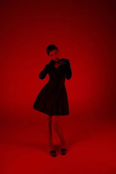 Junges und elegantes asiatisches Model posiert in schwarzem Cocktailkleid, langen Handschuhen und Netzstrumpfhosen auf lebendigem Hintergrund mit rotem Lichteffekt, trendigem Frühling, Mode-Shooting — Stockfoto