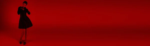 Pleine longueur de jeune femme asiatique en tenue de printemps tendance debout sur fond vibrant avec effet d'éclairage rouge, robe de cocktail noire, gants longs, collants résille, mode jeune, bannière — Photo de stock