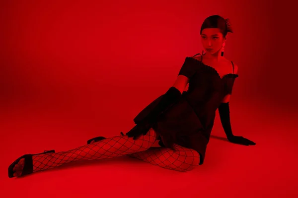 Повна довжина молодої азіатки в стильному весняному вбранні, сидячи і дивлячись на яскравий фон з ефектом червоного освітлення, чорна коктейльна сукня, довгі рукавички, ажурні колготки, мода — стокове фото