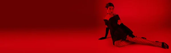 Piena lunghezza della donna asiatica alla moda in abito da cocktail, guanti lunghi neri e calze a rete seduti e guardando lontano su uno sfondo vibrante con effetto luce rossa, concetto di moda primaverile, banner — Foto stock