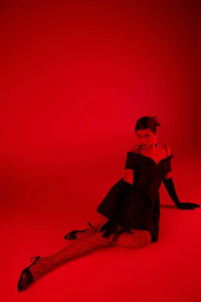 Génération z, concept de mode de printemps, pleine longueur de jeune femme asiatique en robe noire, gants longs et collants résille assis sur un fond vibrant avec effet d'éclairage rouge — Photo de stock