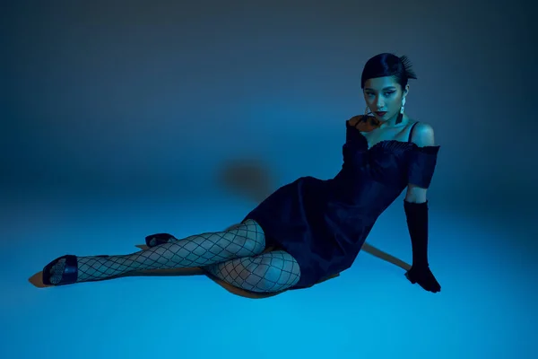 Full length of young and glamour asian fashion model in stylish spring outfit sitting on blue background with cyan lighting effect, black cocktail dress, long glove, fishnet tights — Stock Photo