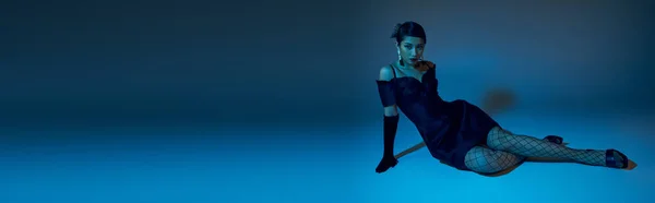 Full length of expressive asian woman in elegant cocktail dress, black long gloves and fishnet tights sitting on blue background with cyan lighting effect, generation z, spring style concept, banner — Stock Photo