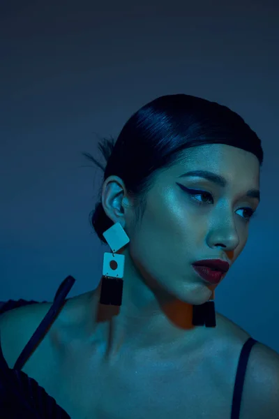 Porträt einer jungen und charmanten Asiatin mit brünetten Haaren, fettem Make-up und trendigen Ohrringen auf blauem Hintergrund mit Cyan-Lichteffekt, stylisches Frühjahrskonzept — Stockfoto