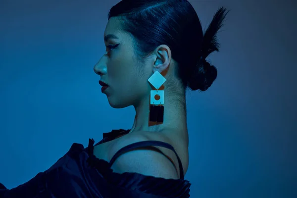 Spring fashion photography, profile asian woman with brunette hair, bold makeup, trendy hairstyle and earring on blue background with cyan lighting effect, generation z, side view — Stock Photo