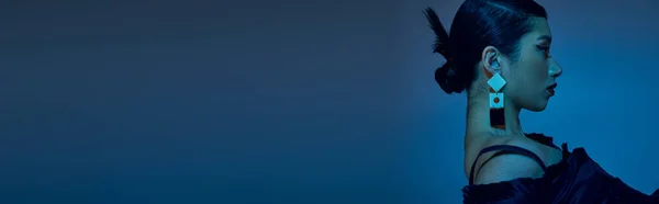 Vista laterale di seducente donna asiatica con capelli castani, orecchino alla moda e trucco audace in posa in abito nero su sfondo blu con effetto luce ciano, concetto alla moda primavera, vista laterale, banner — Foto stock