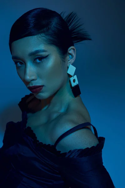 Портрет привлекательной азиатской женщины с брюнетками, смелым макияжем, модной сережкой и выразительным взглядом, смотрящей в камеру на синем фоне с эффектом синего освещения, молодежной культурой, стильной весной — стоковое фото