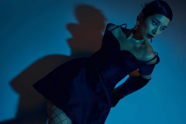 Vista de ángulo alto de la atractiva mujer asiática con cabello moreno, en vestido de cóctel negro y guantes largos mirando hacia otro lado mientras está sentado sobre fondo azul con efecto de iluminación cian - foto de stock