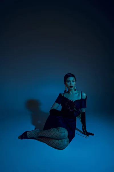 Longitud completa de atractiva mujer asiática en vestido de cóctel negro, medias de rejilla y guantes largos mirando hacia otro lado mientras está sentado sobre fondo azul con efecto de iluminación cyan, modelo de moda - foto de stock