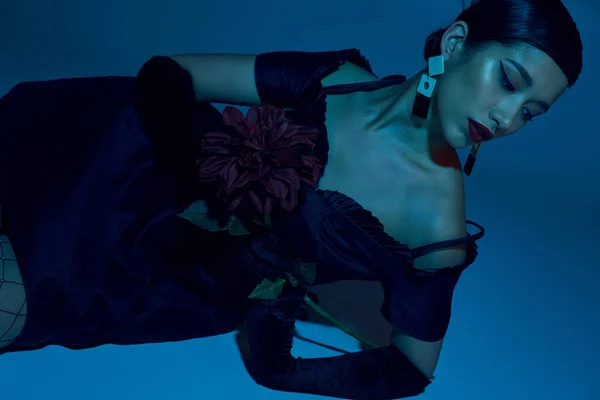 Молодая и чувственная азиатская женщина в модных сережках, черном элегантном платье и длинных перчатках, лежащих с пионскими цветами на синем фоне с эффектом синего освещения, модной весны, модной съемки — стоковое фото