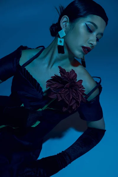 Очаровательная азиатская женщина в черном элегантном платье, длинные перчатки и модные серьги, сидя в стильной позе с цветком пиона на синем фоне с эффектом синего освещения, модная весенняя концепция — стоковое фото