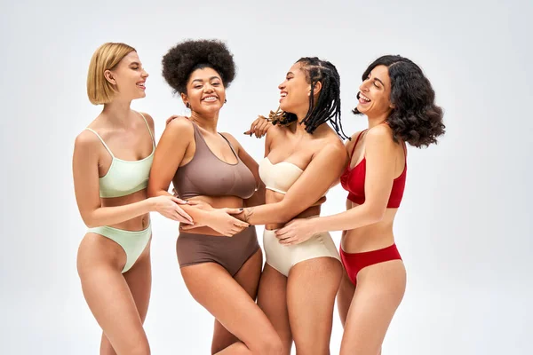 Fröhliche multiethnische Frauen in bunten Dessous umarmen afrikanisch-amerikanische Freundin, während sie isoliert auf grauen, unterschiedlichen Körpertypen und Selbstakzeptanzkonzept stehen und posieren — Stockfoto