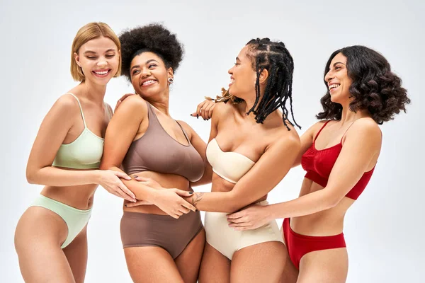 Positive multiethnic women in colorful bras and panties hugging while posing and standing together isolated on grey, different body types and self-acceptance concept, multicultural representation — Stock Photo
