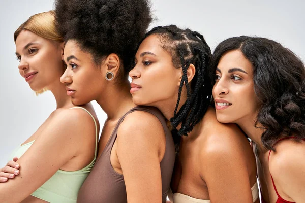 Lächelnde und multiethnische Frauen in bunten BHs, die wegschauen, während sie isoliert nebeneinander auf grauen, unterschiedlichen Körpertypen und Selbstakzeptanzkonzepten stehen, multikulturelle Modelle — Stockfoto