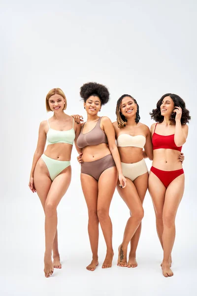 Full length of joyful multiethnic women in colorful bras and panties hugging while standing together on grey background, different body types and self-acceptance concept, multicultural models — Stock Photo
