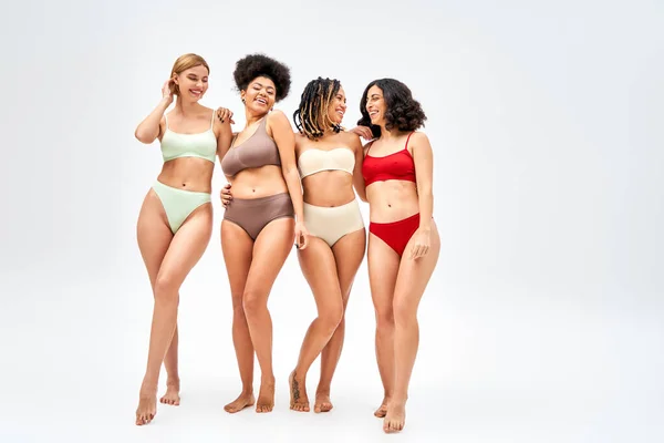 Pleine longueur de heureux groupe multiethnique de femmes en lingerie colorée étreignant et debout sur fond gris, différents types de corps et concept d'acceptation de soi, modèles multiculturels — Photo de stock