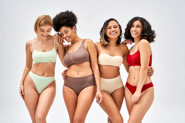 Donne multietniche in lingerie colorata che si abbracciano e guardano la macchina fotografica mentre sono isolate su diversi tipi di corpo grigio e concetto di accettazione di sé, modelli multiculturali — Foto stock