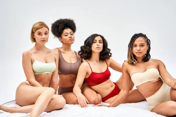 Multiethnic women in colorful bras and panties looking at camera together while sitting on white bed isolated on grey, different body types and self-acceptance concept, multicultural models — Stock Photo