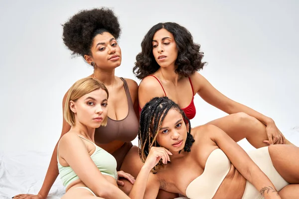 Trendy and multiethnic group of women in colorful lingerie looking at camera while sitting together on white bed isolated on grey, different body types and self-acceptance concept — Stock Photo