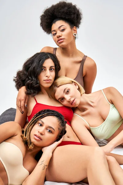 Grupo elegante e multiétnico de mulheres em lingerie colorida diferente olhando para a câmera enquanto posando na cama branca isolada em cinza, diferentes tipos de corpo e conceito de auto-aceitação — Fotografia de Stock