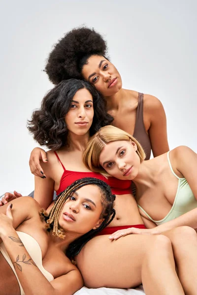 Trendy and multiethnic women in lingerie hugging and looking at camera while sitting on white bed isolated on grey, different body types and self-acceptance concept, multicultural models — Stock Photo