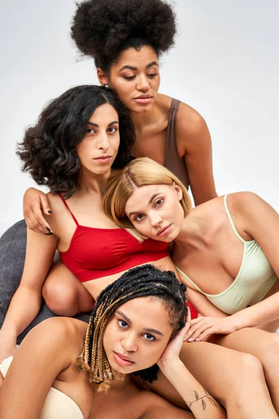 Мультиэтническая группа женщин с различными формами тела в красочном нижнем белье, смотрящих в камеру и обнимающихся, сидя на кровати изолированных на сером, различных типах тела и концепции самопринятия — стоковое фото