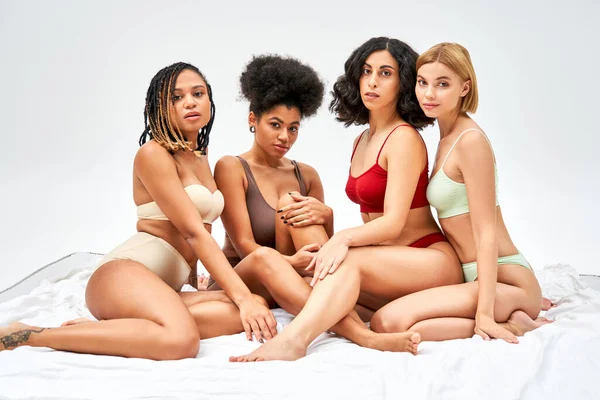 Donne sexy e multietniche in lingerie colorata guardando la fotocamera mentre sedevano insieme su un letto bianco isolato su grigi, diversi tipi di corpo e concetto di accettazione di sé, modelli multiculturali — Foto stock