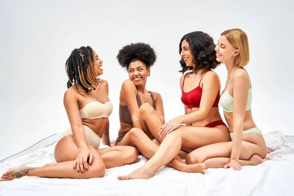 Allegre donne africane americane che parlano vicino ad amici multietnici in lingerie mentre siedono insieme su un letto bianco isolato su grigi, diversi tipi di corpo e concetto di accettazione di sé — Foto stock