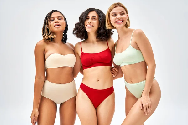 Jovens mulheres multiétnicas alegres em diferentes abraços de lingerie colorida enquanto estão juntas isoladas em cinza, diferentes tipos de corpo e conceito de auto-aceitação, modelos multiculturais — Fotografia de Stock