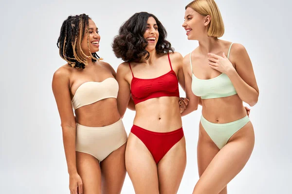Mulheres multiétnicas alegres em lingerie colorida falando ao abraçar e ficar juntos isolados em cinza, diferentes tipos de corpo e conceito de auto-aceitação, modelos multiculturais — Fotografia de Stock