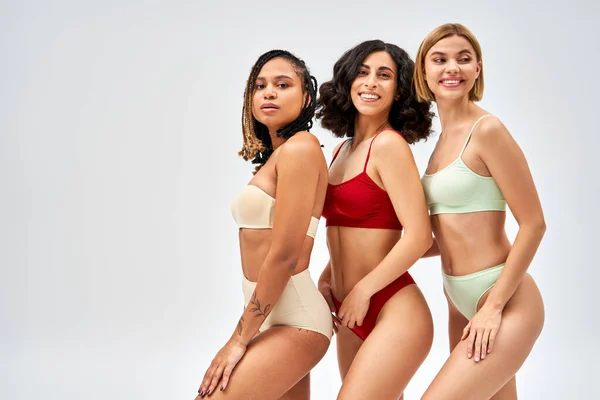 Positive and multiethnic women in stylish colorful lingerie posing while standing next to each other isolated on grey, different body types and self-acceptance concept, multicultural models — Stock Photo
