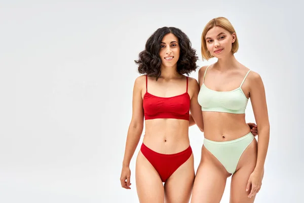 Smiling multiracial woman in lingerie hugging blonde friend and looking at camera while standing isolated on grey, different body types and self-acceptance concept, multicultural models — Stock Photo