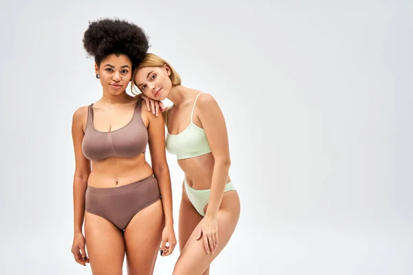 Young blonde woman in trendy lingerie hugging african american friend and looking at camera isolated on grey, different body types and self-acceptance concept, multicultural models — Stock Photo